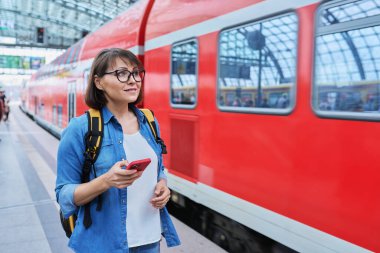 Woman passenger of urban rail transport at modern train station. Female with smartphone, web page with schedule of electric trains, online ticket service, payment for travel through mobile application clipart