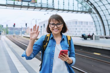 Akıllı telefonlu sırt çantalı, modern tren istasyonu binasının içindeki kameraya bakan olgun, mutlu bir kadın. Duygusal mutluluk, ulaşım, yolculuk, gezi, orta yaşlı insanlar konsepti