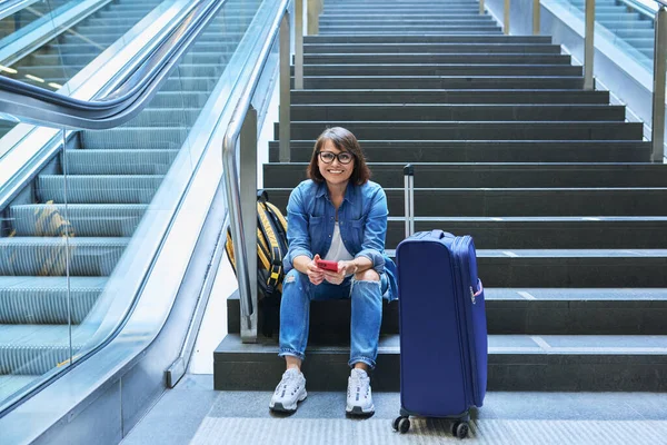 带着行李的女旅客站在车站 中年妇女 手持手提箱智能手机 坐在火车站大楼内的台阶上 运输旅行行李技术人的概念 — 图库照片