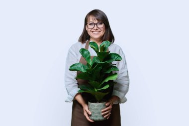 Beyaz stüdyo arkaplanındaki kameraya bakan önlüklü orta yaşlı kadın portresi. Bahçe düzenleme fabrikası. Çiçekçilik reklamı satıyor.