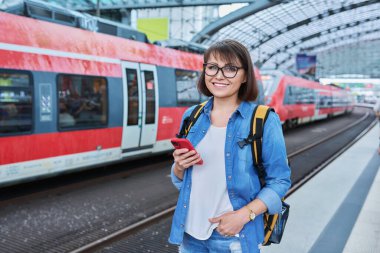 Şehir istasyonunda demiryolu için elektrikli taşımacılık bekleyen bir kadın var. Online bilet ödemesi almak için bankacılık uygulaması kullanan akıllı telefon sahibi bir kadın, çevrimiçi zaman çizelgesi ve rota servisi, teknoloji