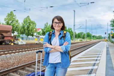 Sırt çantalı, çantalı orta yaşlı bir kadın tren istasyonunun dış peronunda kameraya bakıyor. Ulaşım, bagaj, seyahat, tatil, turizm, insan kavramı