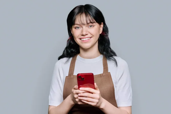 用智能手机笑着穿围裙的年轻女性员工 灰色背景 互联网 咖啡店 小商业概念 — 图库照片