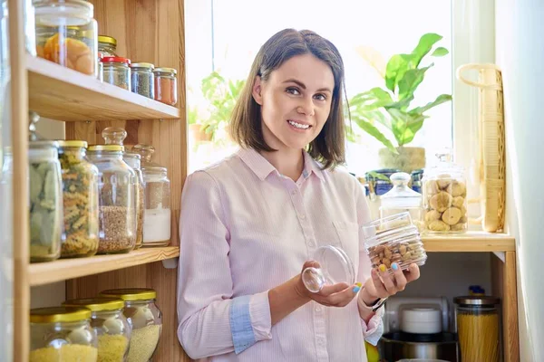 Young woman in kitchen with containers jars of cereals, pasta, dry fruits, holding jar of almonds. Female in home pantry, food organization and storage, kitchen utensils, household