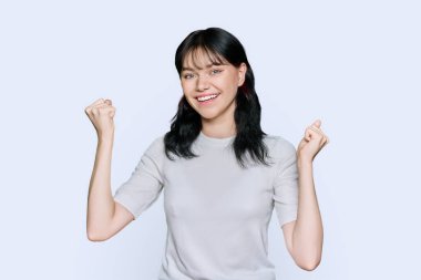 Mutluluk hissi, iyi şans, zafer, kutlama, beyaz stüdyo arka planında ağzı açık, yumruklarını kaldıran genç mutlu kadın. Çekici genç kız öğrenci kameraya bakarak eğleniyor.