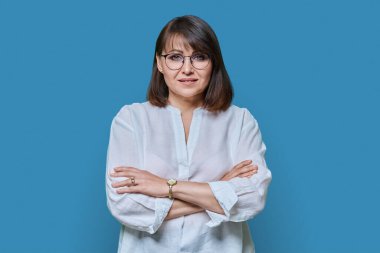 Çekici orta yaşlı kadın mavi arka planda kameraya bakıyor. Gözlüklü, kollarını çapraz beyaz tişörtlü, kendinden emin 40 'lı yaşlarda bir kadın. Yaşam tarzı güven sakinliği olgun insanlar konsepti