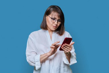 Orta yaşlı odaklı kadın mavi arka planda akıllı telefon kullanıyor. Gerçekten olgun bir kadın telefon ekranında yazı yazıyor. Teknoloji web sitesi mobil uygulamalar yaşam tarzı iş adamları