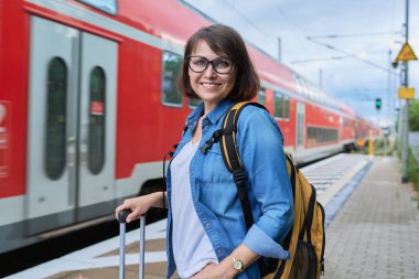 Tren istasyonunun dış peronunda sırt çantalı, orta yaşlı, gülümseyen kadın, kameraya bakıyor, kırmızı elektro trenin arka planına bakıyor. Ulaşım, seyahat, tatil, turizm, insan kavramı