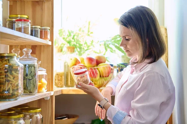 Young woman in kitchen with containers jars of cereals, pasta, dry fruits, holding jar of spices ground red pepper. Female in home pantry, food organization and storage, kitchen utensils, household