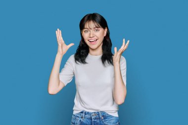 Mutluluk hissi, iyi şans, zafer, kutlama, mavi arka planda ağzı açık, el ele tutuşan mutlu genç bir kadın. Çekici genç kız öğrenci kameraya bakarak eğleniyor.