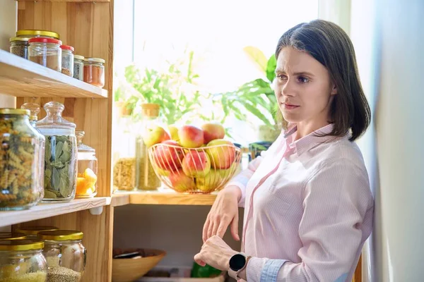 Young woman in kitchen near wooden rack with cereal products, jars of food containers. Pantry, food stock, kitchen utensils, cooking at home, organizing storage concept