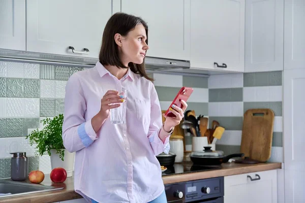 Young woman at home in kitchen looking in smartphone, with glass of water in hands. Beautiful smiling female drinking water, resting, using mobile apps, home interior background