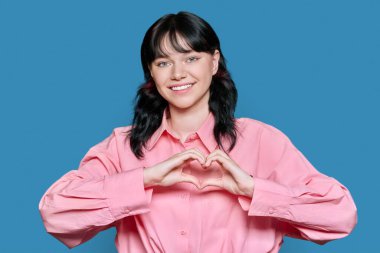 Genç, gülümseyen bir kadın kalp hareketini parmaklarıyla gösteriyor, mutlu bir kadın sevgisini gösteriyor, mavi bir geçmişi var. Semboller, aşk, duygular, romantizm gençlik kavramı