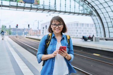 Akıllı telefonlu sırt çantalı, modern tren istasyonu binasının içindeki kameraya bakan olgun, mutlu bir kadın. Duygusal mutluluk, ulaşım, yolculuk, gezi, orta yaşlı insanlar konsepti