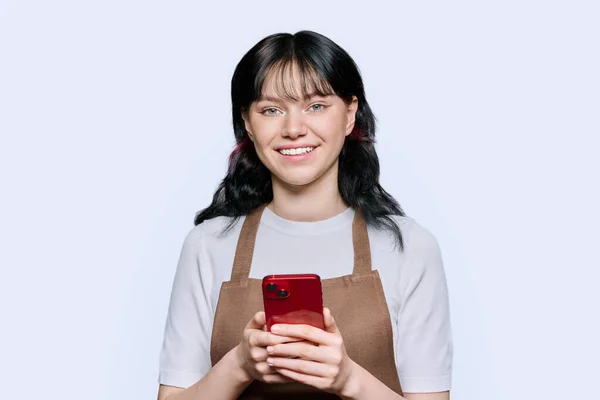 用智能手机笑着穿围裙的年轻女性员工 在白色的工作室背景下看着相机 互联网 咖啡店 小商业概念 — 图库照片