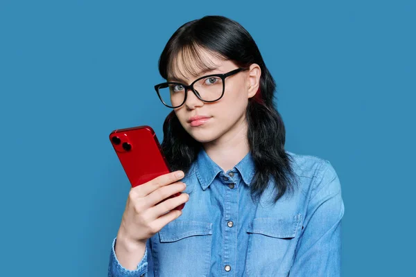 戴眼镜 手持智能手机 拿着蓝色摄影棚背景相机的认真的女学生 — 图库照片