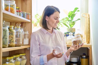 Mutfakta elinde mısır gevreği kavanozları, makarna, kuru meyve ve elinde bir kavanoz bademle genç bir kadın var. Kilerde kadın, yemek organizasyonu ve depolama, mutfak aletleri, ev eşyaları.