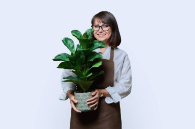 Beyaz stüdyo arkaplanındaki kameraya bakan önlüklü orta yaşlı kadın portresi. Bahçe düzenleme fabrikası. Çiçekçilik reklamı satıyor.