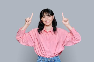 Genç bir kadın parmaklarını havaya kaldırdı. Gri arkaplanda, fotokopi için yer açtı. Kameraya bakan çekici bir genç kız. Reklamcılık, pazarlama konsepti