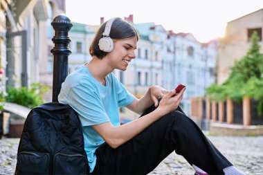 Akıllı telefondan video görüşmesi yapan yakışıklı bir adam. Şehir caddesinde açık havada. Kulaklık takan, sırt çantalı, internette konuşan genç bir erkek. Görüntülü sohbet, iletişim teknolojisi.
