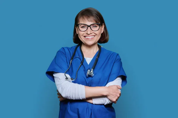 带着听诊器微笑的中年医生的画像 在蓝色的工作室背景上看着相机 戴眼镜 两臂交叉的自信女人 医疗护理人员医疗服务概念 — 图库照片