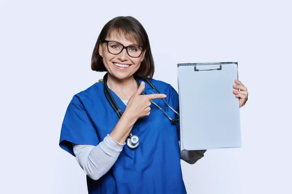 女医生指指剪贴板上的空白纸 白色工作室背景 穿着蓝色制服的友善的中年护士 剪贴板空间 用于个人留言建议 广告文字 — 图库照片