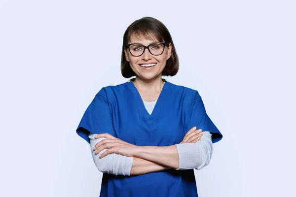 身着蓝色制服 面带微笑的中年护士的画像 在白色的工作室背景上看着相机 戴眼镜 双臂交叉的自信积极的女人 医疗护理人员医疗服务 — 图库照片