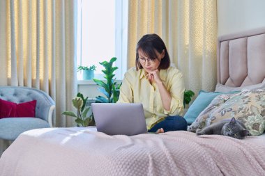 Orta yaşlı bir kadın evinde, kanepede dizüstü bilgisayarı ve kedisiyle oturuyor. Olgun 40 'lı yaşlarda bir kadın evcil hayvanla yatakta ekrana bakıyor. Evde çalışmak, teknoloji, eğlence, serbest çalışmak, yaşam tarzı, insanlar ve hayvanlar konsepti