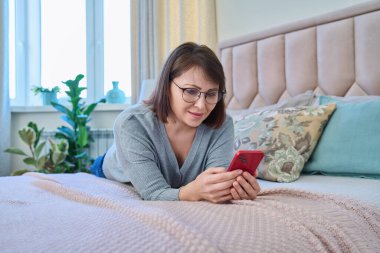 Middle aged relaxed woman lying on bed at home using smartphone mobile apps for leisure leisure reading watching video. Lifestyle, technology, 40s people concept