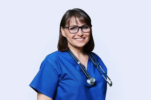 身着蓝色制服的中年女护士 带着听诊器微笑看着相机 在白色的工作室背景上刻画成熟女医生的肖像 医务人员概念 — 图库照片