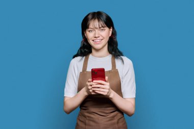 Önlüklü gülümseyen genç bayan çalışanlar akıllı telefon kullanıyor, mavi arkaplandaki kameraya bakıyor. Servis, internet, başvuru, personel, kafe, dükkan, dükkan, küçük işletme konsepti