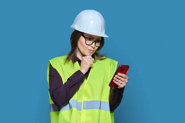 女性工业物流建筑工人穿着带智能手机的头盔背心 用手机应用于蓝色背景的工作 互联网技术建筑业管理工程人员 — 图库照片