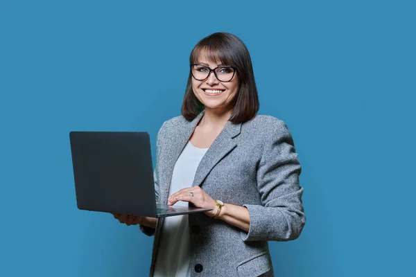 Middle age business woman with laptop looking at camera on blue color background. Positive successful mature female using laptop for business, service. Entrepreneurship finance employment study