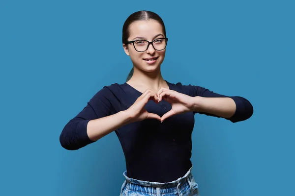 Young smiling female showing heart gesture with fingers, happy female showing love, blue studio background. Body language, signs symbols, love, emotions, romance youth concept