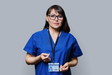 Orta yaşlı kadın tıp merkezi çalışanı gri stüdyo arka planındaki kameraya bakıyor..