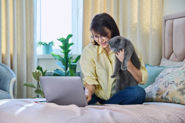 Middle aged woman at home on couch with laptop and cat. Mature 40s female looking at screen, on bed with pet. Work at home, technology, leisure, freelancing, lifestyle, people and animals concept