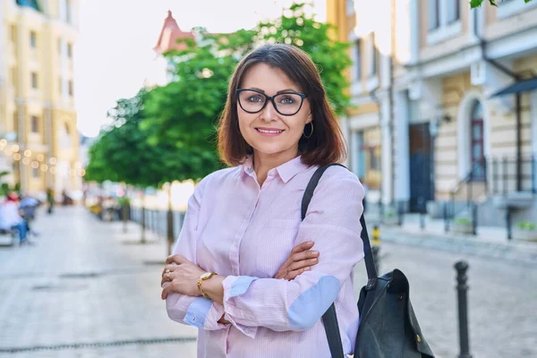 Portrait of positive confident middle aged woman looking at camera outdoor. Smiling female in glasses, arms crossed on city street. Business, age, positive, confidence, 40s people concept