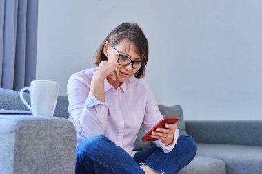 Orta yaşlı, rahat, gülümseyen bir kadın evinde koltukta oturuyor, akıllı telefon kullanıyor, kısa mesajlar okuyor, boş zamanlarında cep telefonu uygulamaları kullanıyor.