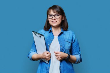 Mavi arka plandaki kameraya bakan orta yaşlı, kendine güvenen bir iş kadını. Olgun gülümseyen bayan öğretmen ajan müfettiş psikolog evraklarla poz veriyor.