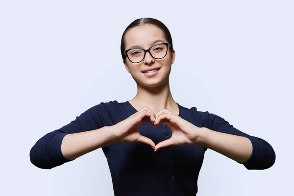 Young smiling female showing heart gesture with fingers, happy female showing love, white studio background. Body language, signs symbols, love, emotions, romance youth concept