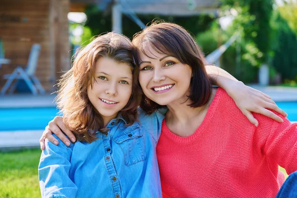Portrait of happy mom and teenage daughter looking at camera. Smiling hugging mother and girl together in backyard near outdoor pool. Family, parent and child teenager, mothers day, lifestyle concept