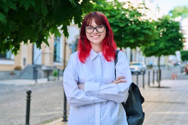 Sırt çantasıyla kameraya bakan, gözlüklü, kendine güvenen genç bir bayan. Şehirde kızıl boyalı saçlı, güzel, modaya uygun bir kız. Güzellik, gençlik, yaşam tarzı konsepti