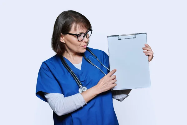 女医生指指剪贴板上的空白纸 白色工作室背景 严肃的中年护士 身穿蓝色制服 剪贴板空间个人留言建议信息广告文字 — 图库照片