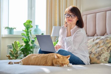Orta yaşlı bir kadın evinde, kanepede dizüstü bilgisayarı ve kedisiyle oturuyor. Olgun 40 'lı yaşlarda bir kadın kameraya bakıyor, evcil hayvanla yatakta. Evde çalışmak, teknoloji, eğlence, serbest çalışmak, yaşam tarzı, insanlar ve hayvanlar konsepti