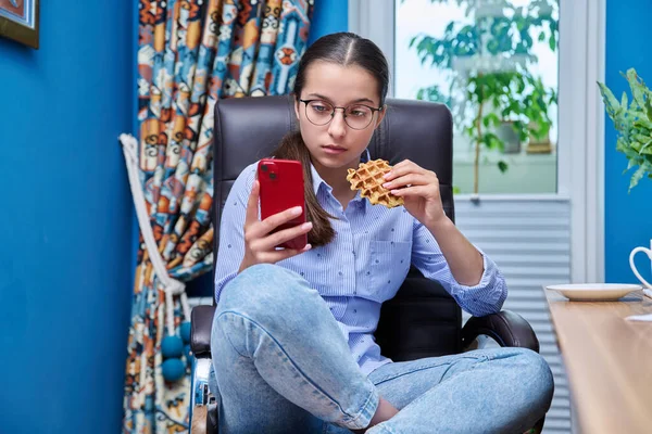 소녀가 컴퓨터 근처의 의자에 스마트폰을 와플을 있습니다 암컷은 집에서 공부하며 — 스톡 사진