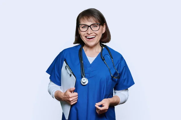 女医生形象与剪贴板看着相机在白色工作室背景 中年医生护士 带听诊器 身穿蓝色制服 工作人员概念 — 图库照片