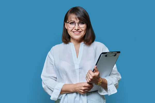 Confident middle aged business woman with clipboard looking at camera on blue background. Mature smiling female teacher agent auditor psychologist supervisor posing with papers documents contracts