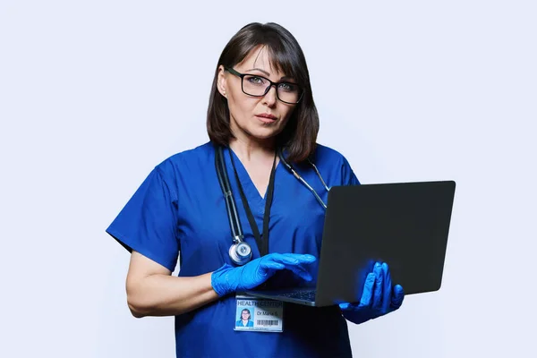 Portrait of serious female doctor with laptop, confident female looking at camera on white studio background. Medic nurse with stethoscope in blue uniform. Health care medicine internet service staff