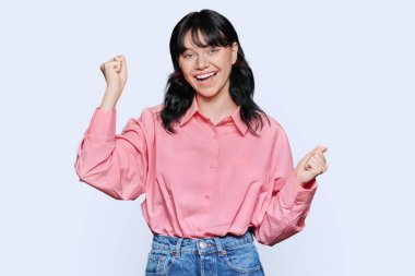 Mutluluk hissi, iyi şans, zafer, kutlama, beyaz stüdyo arka planında ağzı açık, yumruklarını kaldıran genç mutlu kadın. Çekici genç kız öğrenci kameraya bakarak eğleniyor.