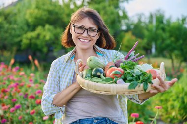 Portrait of smiling middle aged woman with basket of different fresh raw vegetables and herbs, summer nature vegetable garden background. Harvest from organic farm, healthy food nutrition, gardening